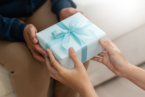 Close up on hands, Daughter is giving a blue gift box to her mother while her sitting on couch for New year, Birthday, Christmas. Celebration, Anniversary.
