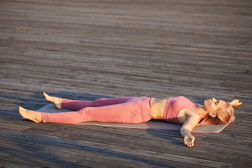 Young slim woman in pink sports clothing lying on exercise mat and relaxing with her eyes closed outdoors