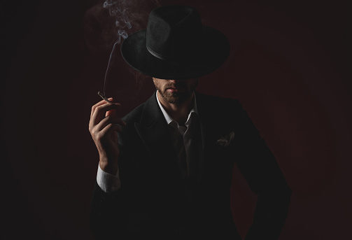 mysterious young man wearing tuxedo and black hat, smoking cigarette and looking down, sitting on black background