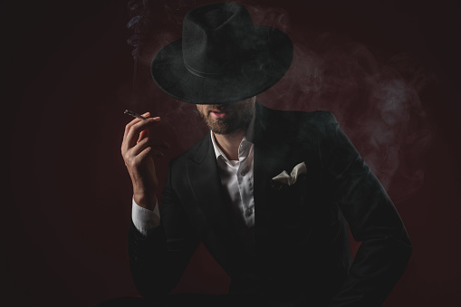 attractive young man wearing tuxedo and black hat, looking down, smoking cigarette and sitting on black background