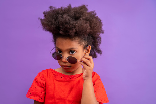 Ethnic kid girl looking camera lowering sunglasses on purple background
