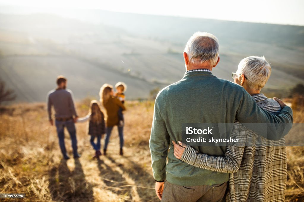 Rear view of embraced senior couple looking at their family in nature. Back view of embraced grandparents enjoying while looking at their family on a field in autumn day. Family Stock Photo