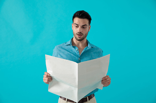 attractive casual man in blue shirt is standing and reading the newspaper shocked on blue studio background