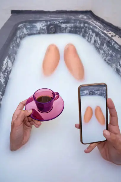 woman hand drinking coffee pink cup taking selfie photo of her legs at milk bathtub