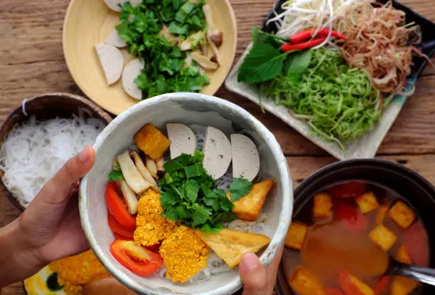 Top view woman hand prepare homemade breakfast, vegan rieu noodle soup or vegetarian crab paste vermicelli soup, a traditional Vietnamese dish, plate of vegetables, herbs, soup pot, ready to eat