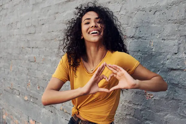 Cropped portrait of an attractive young woman standing alone outside and making a heart sign gesture against a gray background