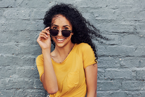 Cropped portrait of an attractive young woman wearing sunglasses and smiling while standing against a gray background outdoors