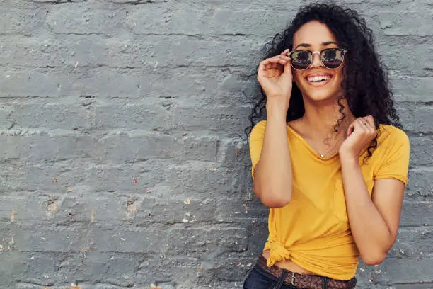 Cropped portrait of an attractive young woman wearing sunglasses and smiling while standing against a gray background outdoors