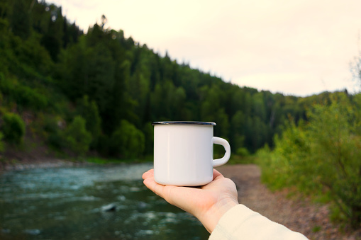 Woman holding a white campfire enamel mug mockup with riverside view. Empty mug mock up for design promotion.