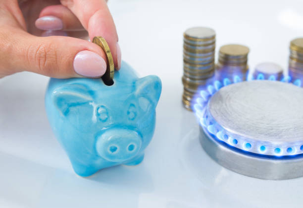 Woman's hand puts a coin in a piggy bank near the blue flame of domestic gas in the kitchen stove. The symbolic image of saving natural gas, paying for gas consumed, energy efficiency at home. stock photo