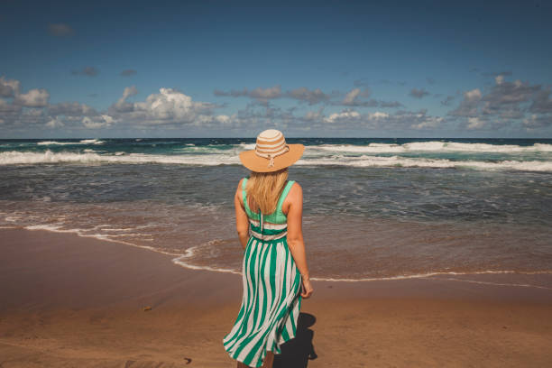 View from behind of a young woman looking out to sea and waves from a sandy beach on a sunny day.