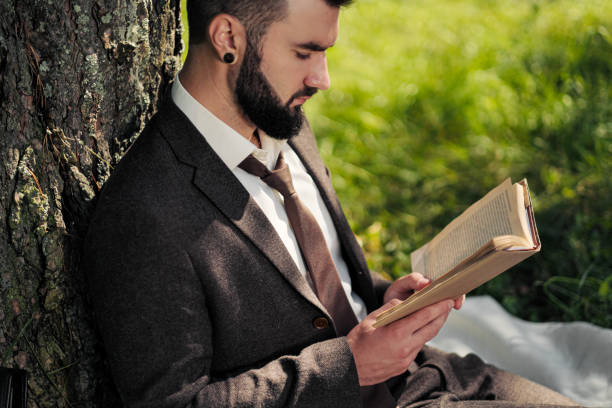 istock Young attractive bearded business man sitting on green grass under tree and resting in park. Read book, drink coffee. Relaxed, tired of work, lunch break. 1175009907