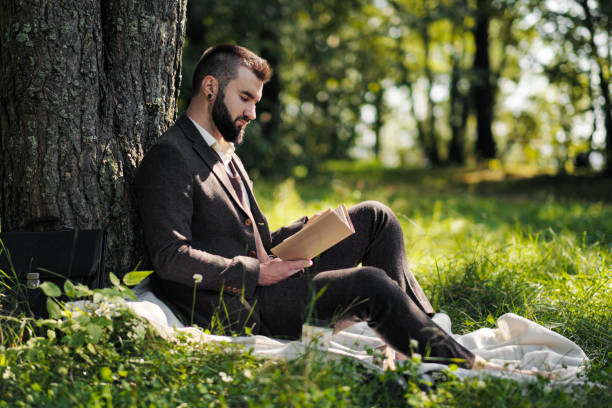 istock Young attractive bearded business man sitting on green grass under tree and resting in park. Read book, drink coffee. Relaxed, tired of work, lunch break. 1175009909