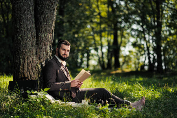 istock Young attractive bearded business man sitting on green grass under tree and resting in park. Read book, drink coffee. Relaxed, tired of work, lunch break. 1175009921