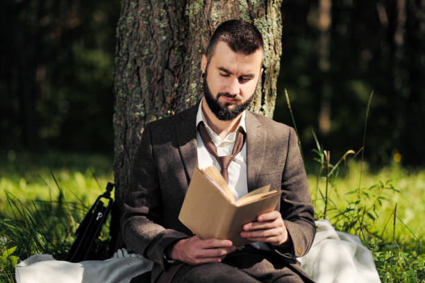 istock Young attractive bearded business man sitting on green grass under tree and resting in park. Read book, drink coffee. Relaxed, tired of work, lunch break. 1175009933