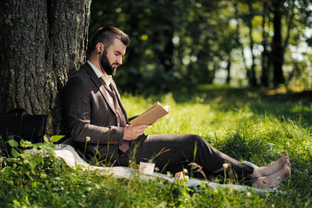 istock Young attractive bearded business man sitting on green grass under tree and resting in park. Read book, drink coffee. Relaxed, tired of work, lunch break. 1175009979