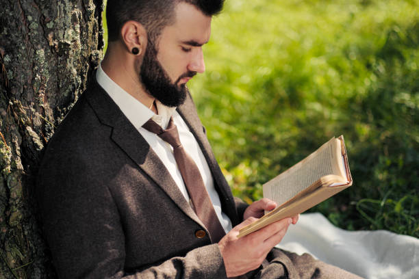 istock Young attractive bearded business man sitting on green grass under tree and resting in park. Read book, drink coffee. Relaxed, tired of work, lunch break. 1175009989