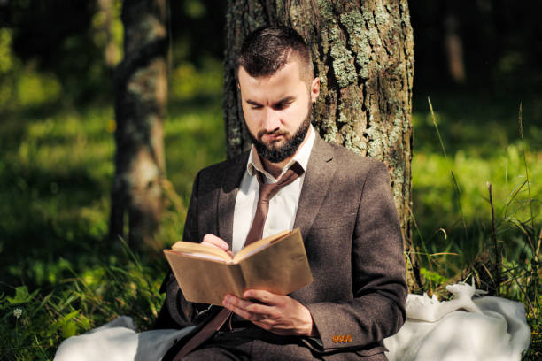 istock Young attractive bearded business man sitting on green grass under tree and resting in park. Read book, drink coffee. Relaxed, tired of work, lunch break. 1175009990