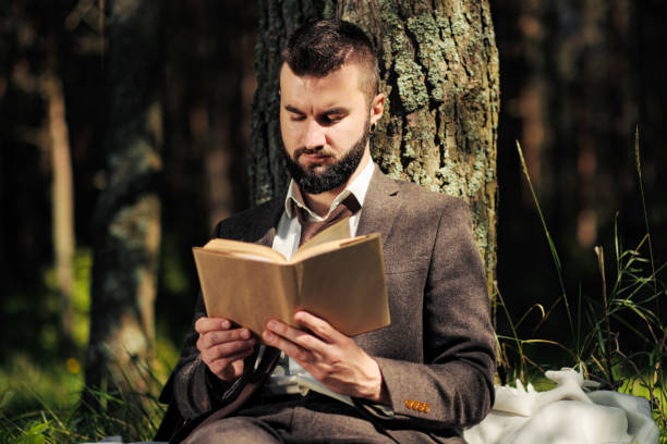Young attractive bearded business man sitting on green grass under tree and resting in park. Read book, drink coffee. Relaxed, tired of work, lunch break.