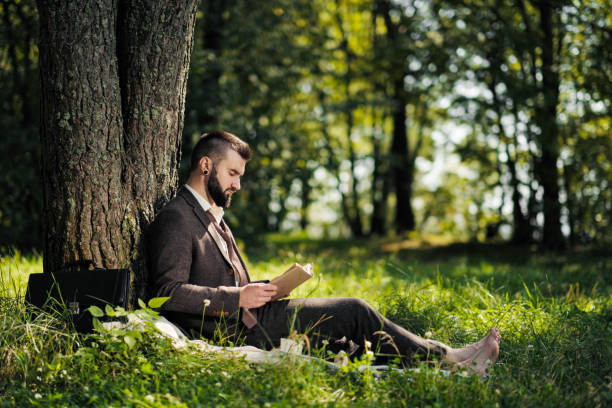 istock Young attractive bearded business man sitting on green grass under tree and resting in park. Read book, drink coffee. Relaxed, tired of work, lunch break. 1175010038