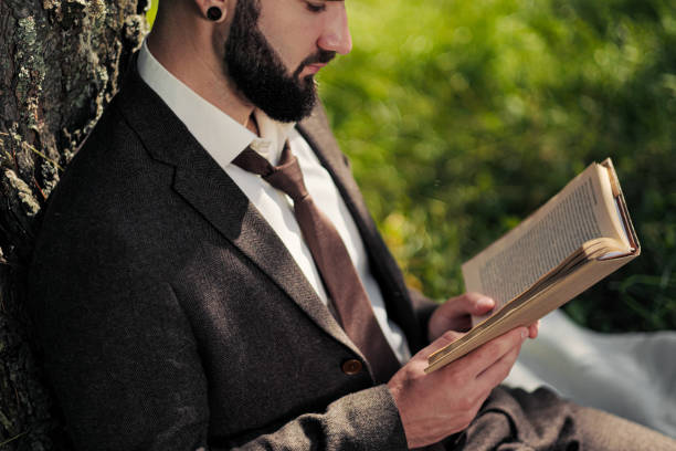istock Young attractive bearded business man sitting on green grass under tree and resting in park. Read book, drink coffee. Relaxed, tired of work, lunch break. 1175010055