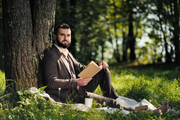 istock Young attractive bearded business man sitting on green grass under tree and resting in park. Read book, drink coffee. Relaxed, tired of work, lunch break. 1175010111