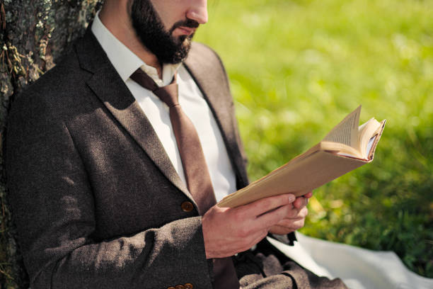 istock Young attractive bearded business man sitting on green grass under tree and resting in park. Read book, drink coffee. Relaxed, tired of work, lunch break. 1175010114