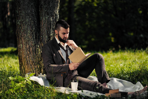 istock Young attractive bearded business man sitting on green grass under tree and resting in park. Read book, drink coffee. Relaxed, tired of work, lunch break. 1175011002
