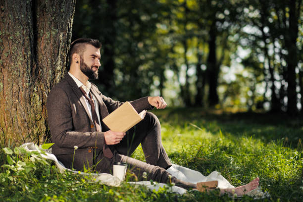 istock Young attractive bearded business man sitting on green grass under tree and resting in park. Read book, drink coffee. Relaxed, tired of work, lunch break. 1175011045