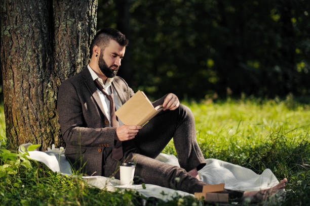 istock Young attractive bearded business man sitting on green grass under tree and resting in park. Read book, drink coffee. Relaxed, tired of work, lunch break. 1175011144