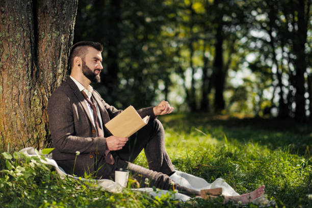 istock Young attractive bearded business man sitting on green grass under tree and resting in park. Read book, drink coffee. Relaxed, tired of work, lunch break. 1175011188