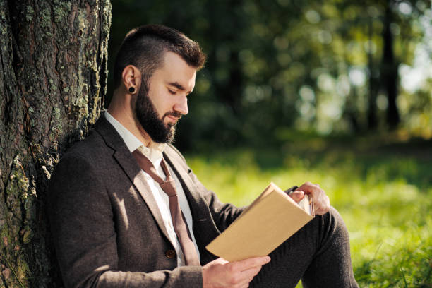 istock Young attractive bearded business man sitting on green grass under tree and resting in park. Read book, drink coffee. Relaxed, tired of work, lunch break. 1175011271