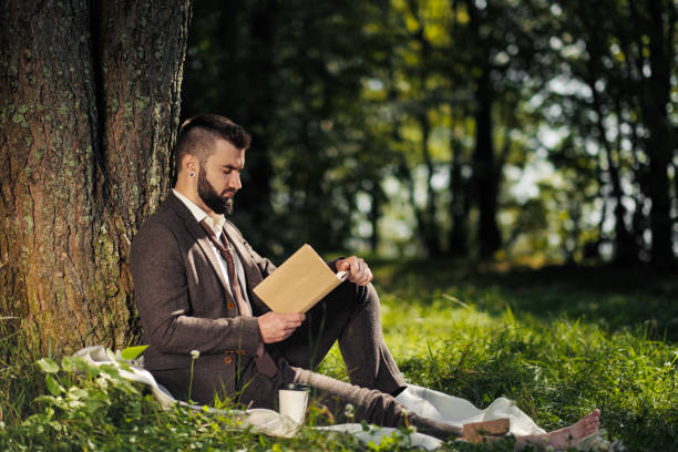 istock Young attractive bearded business man sitting on green grass under tree and resting in park. Read book, drink coffee. Relaxed, tired of work, lunch break. 1175011375