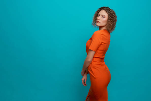 Side view of beautiful, stylish model in orange dress standing on blue background with copy space. Elegant, young woman with curly hair, perfect skin, make up looking at camera.