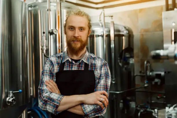 A portrait of handsome brewer in uniform at the beer manufacture with metal containers on the background, who is making beer on his workplace in the brew-house