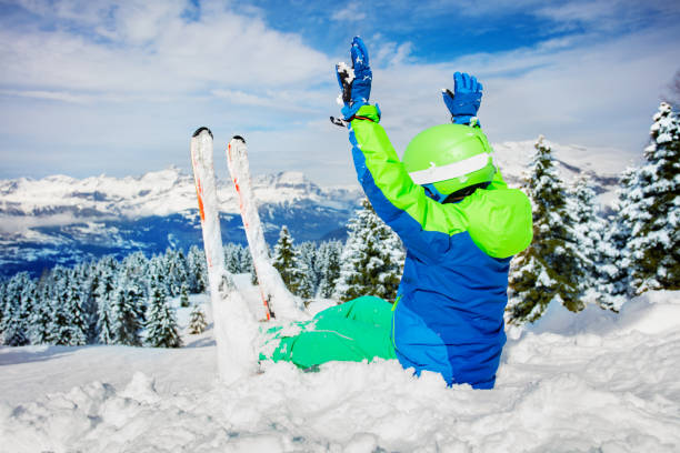 Little boy sit in deep snow with ski over mountain range and snowy forest lifting hands