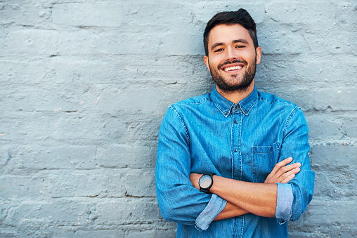 Cropped portrait of a handsome young man smiling while standing with his arms crossed against a brick wall