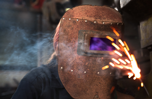 Female mechanical engineer welding metal in factory workshop wearing protective safety helmet - Apprentice industrial student using production equipment - training, industry and construction concept