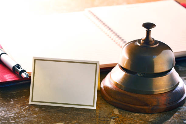 A blank card rests next to a service bell on a hotel reception desk.  An open reservation book and a fountain pen sit out of focus in the background.