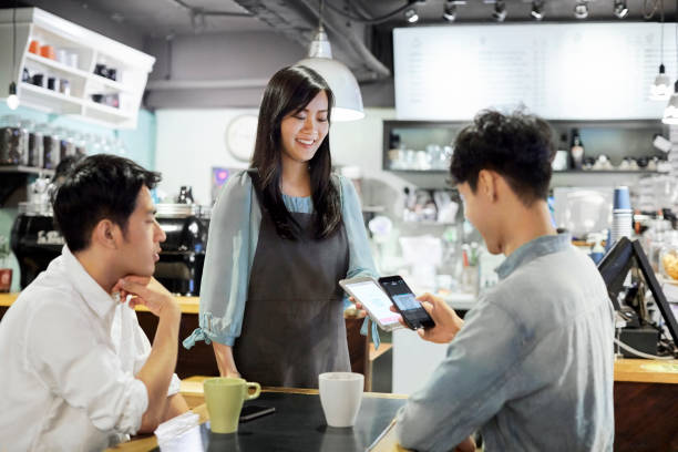 Male customer doing contactless payment to barista Male customer paying through mobile phone while sitting with friend. Young men are doing contactless payment to female barista. They are at cafeteria. point of sale tablet stock pictures, royalty-free photos & images