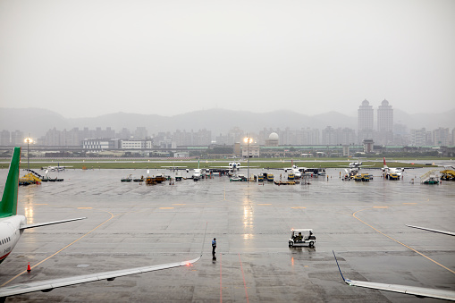 High angle view of wet airport runway. Distant view of airplanes parked in city during rainy season. Cityscape and mountains in foggy weather against sky.