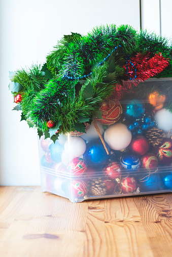 Storage of Christmas decorations. Christmas balls, decorations, tinsel and garlands in a large plastic box on a wooden table, light background.
