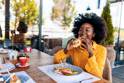 Happy young African woman eating sandwich and smiling while sitting in the restaurant during the day. Taking break.