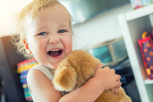 Cute adorable caucasian blond toddler boy having fun, laughing and hugging soft puppy toy indoors. Cheerful child playing with teddy bear at home. Kid friend animal toy. Happy childhood concept.