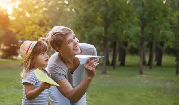 Photo of grandmother and child granddaughter are playing with paper planes and smiling while spending time outdoors