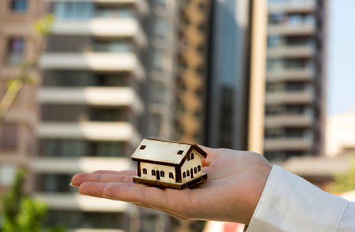Woman hands holding a model house. City district, buildings and blue sky in background. New home, buying and renting, mortgage, real estate concepts. Horizontal close-up detail.