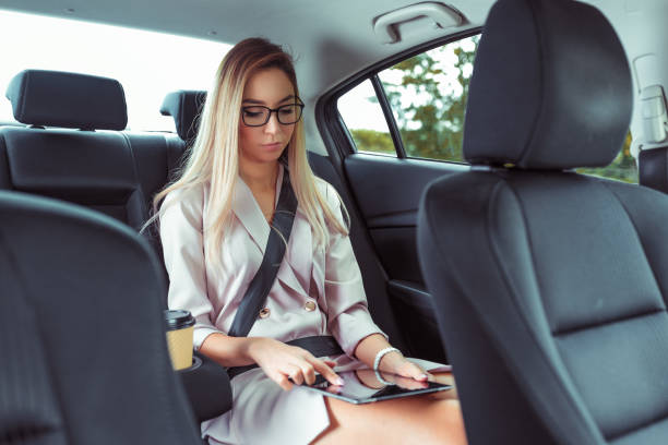 Business woman in suit, sits in VIP taxi, holds tablet in hands, presses finger on touch screen, cup of coffee tea, in summer city, beautiful woman in glasses secretary works on road. stock photo