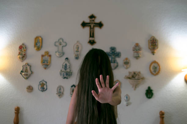 Young girl with long mane in front of her face sitting on the bed shows the palm of her hand in a dark room, in the background there is a cross and several religious elements Young girl with long mane in front of her face sitting on the bed shows the palm of her hand in a dark room, in the background there is a cross and several religious elements exorcism stock pictures, royalty-free photos & images