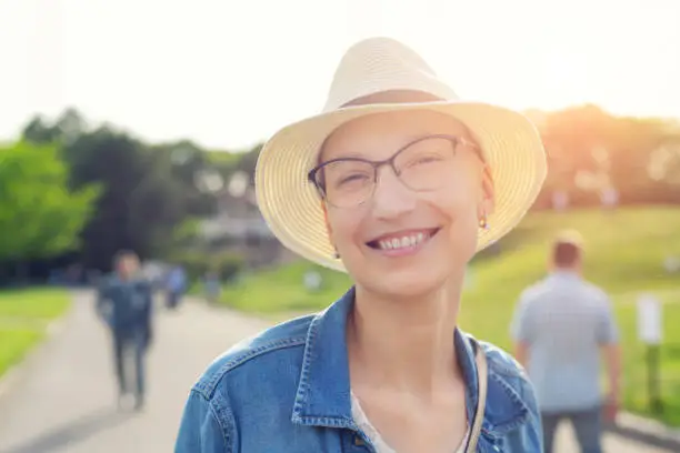 Photo of Happy young caucasian bald woman in hat and casual clothes enjoying life after surviving breast cancer. Portrait of beautiful hairless girl smiling during walk at city park after curing disease