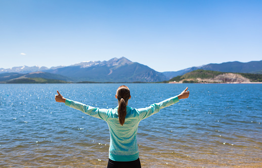 Young female standing next to mountain lake with arms up feeling adventurous happy and free. Location Colorado mountains.